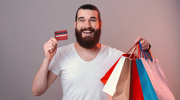 用信用卡在网上购物，快乐的大胡子年轻人拿着纸购物袋和塑料卡。图片下载