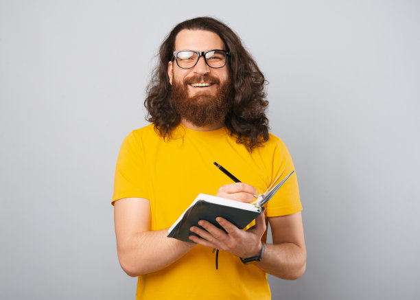 照片中，一个快乐的年轻人，留着长发，微笑着在议程上做笔记。图片下载