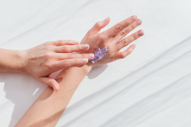 女性手在皮肤上应用紫色霜磨砂膏俯视图白色背景。护肤程序的概念图片下载