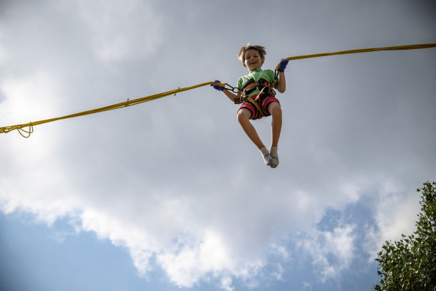 那个男孩正在蹦极蹦床上跳。一个带着保险和可伸缩橡皮筋的孩子悬挂在天空中。快乐童年的概念和游乐园的游戏图片下载