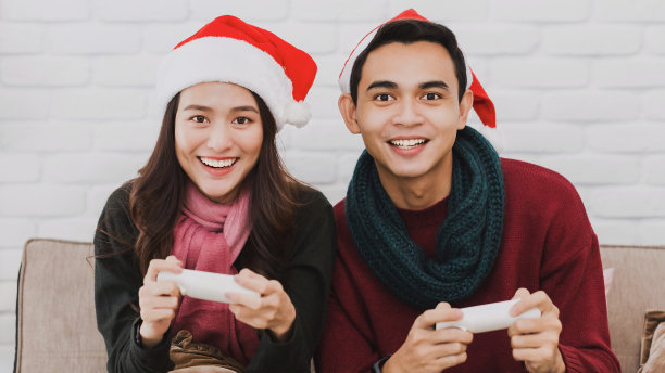 年轻的亚洲情侣戴着圣诞老人的帽子。在装饰着圣诞树的房间里一起玩电子游戏。圣诞节庆祝活动的概念。图片下载