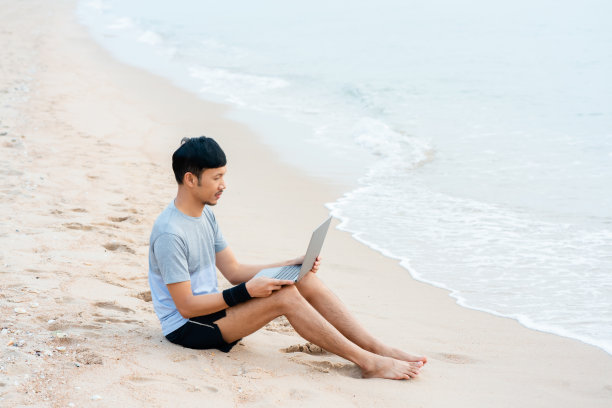 在海边，凉爽的海浪拍打着他，英俊的年轻亚洲男子在他的笔记本电脑上与主管讨论工作。图片下载