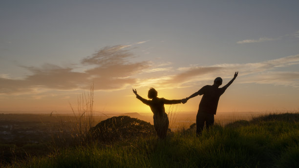 一对剪影夫妻站在山顶上，张开双臂迎接太阳。奥克兰，树山一家。图片下载