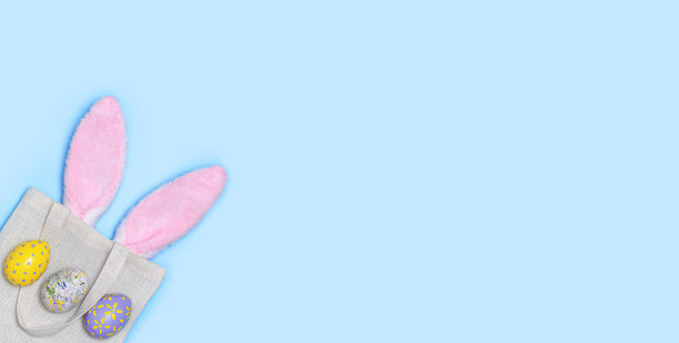 Banner：粉色复活节兔子耳朵从可重复使用的纺织袋中伸出，蓝色背景上画着彩蛋图片下载