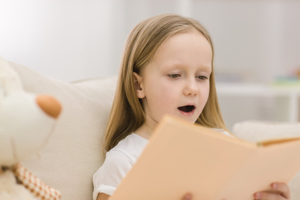 金发小女孩坐在沙发上看书的照片。女孩拿着书的概念。图片下载