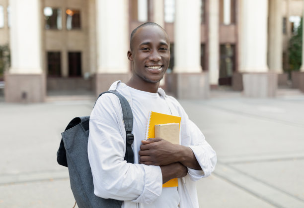 奖学金资助。黑人男学生的肖像，背包和书摆姿势在大学建筑的背景下图片下载