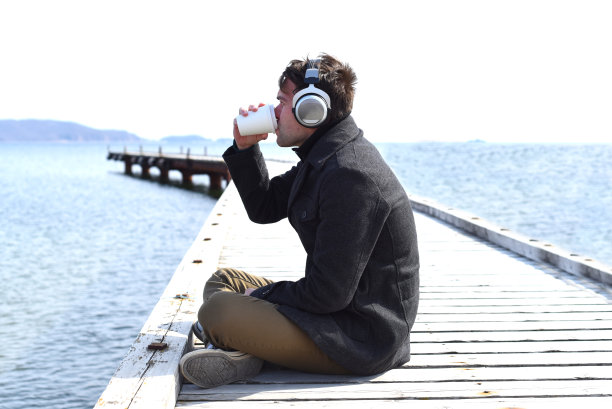 年轻人听着音乐，喝着咖啡，在阳光明媚的日子里独自坐在码头上图片下载