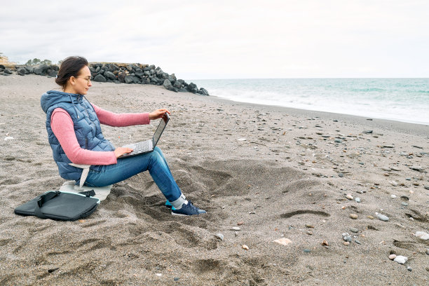 美丽的运动妇女戴着眼镜，笔记本电脑户外工作，而坐在海边的冬季海滩。现代生活方式，人际关系，博客，商业，自由职业。图片下载