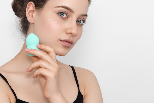 一个年轻的女孩拿着一块用来化妆的蓝绿色海绵看着镜头，脸上涂着粉底。图片下载