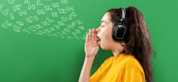 在线语言学习。一个戴着耳机的年轻女子的肖像，她的手捂着她张开的嘴。绿色背景与字母。侧视图。横幅。英语日的概念图片下载