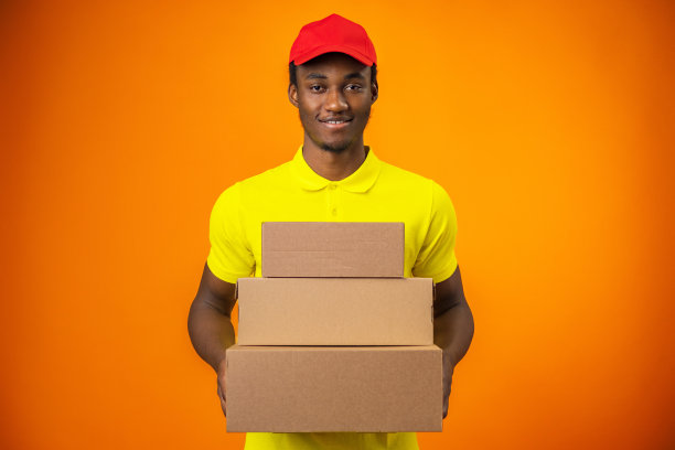 微笑的黑人快递员穿着制服，拿着盒子在橙色的工作室里图片下载
