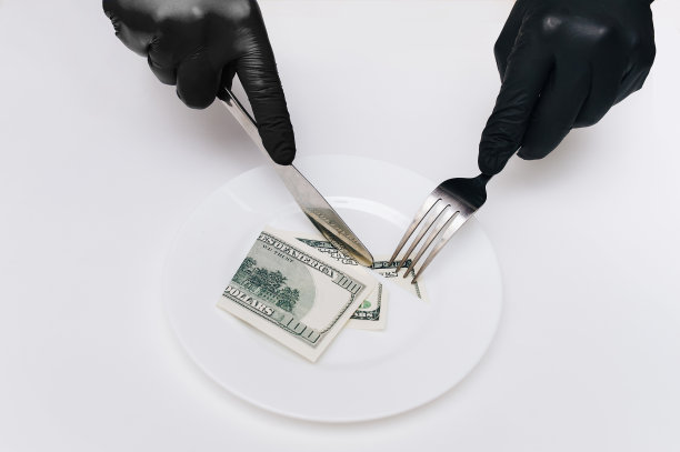 商务午餐的概念。戴着黑色手套的女性手拿着刀叉，从一张百元大钞上切下一块放在一个白色盘子上。设想可视化税收，贬值和利息。图片下载