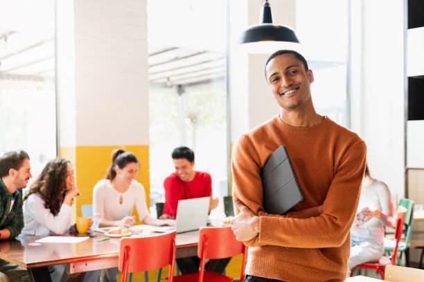 穿着休闲服装的非洲青年拿着写字板，微笑着站在镜头前。一群在后面工作的人图片下载
