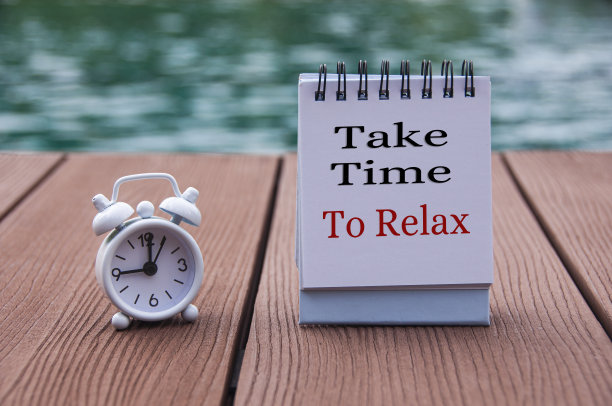 木地板上的闹钟和记事本上的文字——花点时间放松一下。与模糊的游泳池背景。钟定在九点钟。图片下载