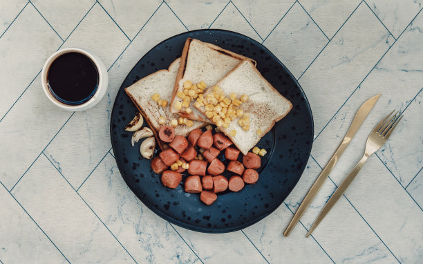 早餐有炸香肠、面包、甜玉米粒和大蒜，用黑色陶瓷盘子盛着，配上一杯黑咖啡。图片下载