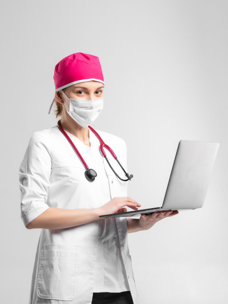漂亮的白种人年轻医生，穿着白色的医疗制服，戴着防毒面具，在工作室里用笔记本电脑。概念照片在工作室，灰色背景。图片下载