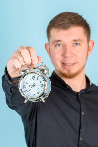 一个留着胡子的年轻人拿着一个闹钟站在蓝色背景上，选择性地聚焦图片下载