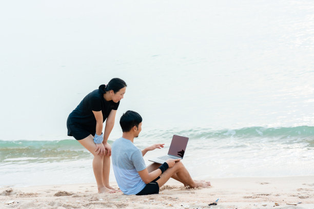 这对年轻夫妇在沙滩上用笔记本电脑通过视频和同事聊天，那里有缓慢的海浪拍打着海岸，快乐而舒适。图片下载