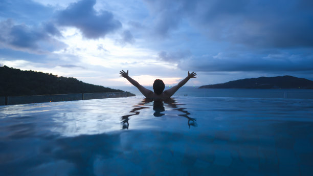 后视图亚洲人伸展手臂胜利姿势放松在户外泳池度假海景日落天空。图片下载