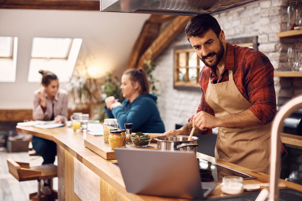 一个年轻人在厨房用笔记本电脑为他的朋友做饭。图片下载