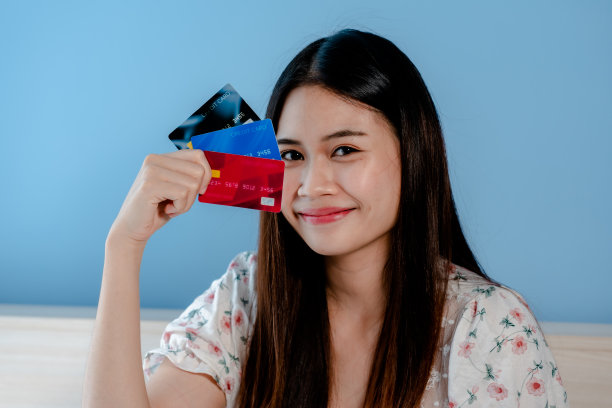 一个美丽的亚洲女孩，坐在床上，穿着白色花衬衫，正用她的手机在网上订购，表情严肃，在她的手机里有三张信用卡，图片下载