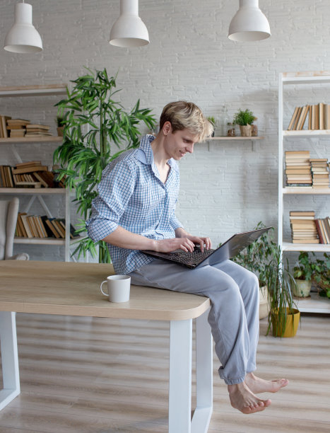 一个年轻人正坐在桌子上，双腿垂下，在笔记本电脑上工作。图片下载