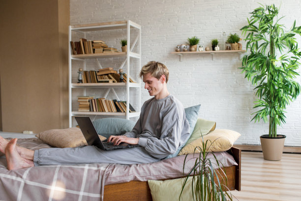 20岁的年轻英俊男子，在笔记本电脑上工作，躺在床上休息，在家里的卧室里消磨时间。图片下载