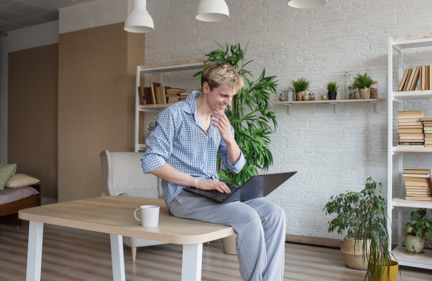 一个年轻人正坐在桌子上，双腿垂下，在笔记本电脑上工作。图片下载