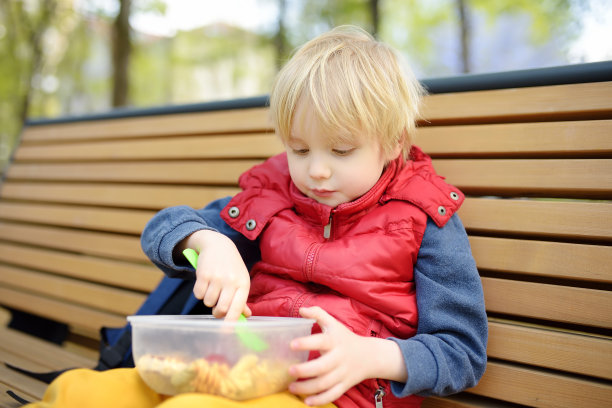 幼儿园或学校放学后，一个小男孩坐在公园长凳上的塑料容器里吃午饭。在街上为孩子们带走食物。给孩子们的健康食品。图片下载