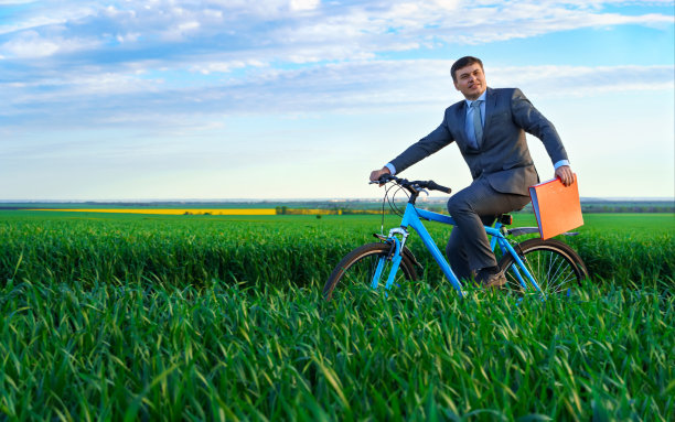 一个商人拿着红色的报告或文件文件夹骑着自行车穿过一片绿色的草地，穿着西装，在美丽的大自然夏天，经营理念图片下载