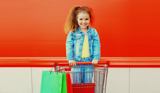 快乐微笑的小女孩的肖像，带着手推车和购物袋，穿着牛仔布的衣服在城市的街道上图片下载
