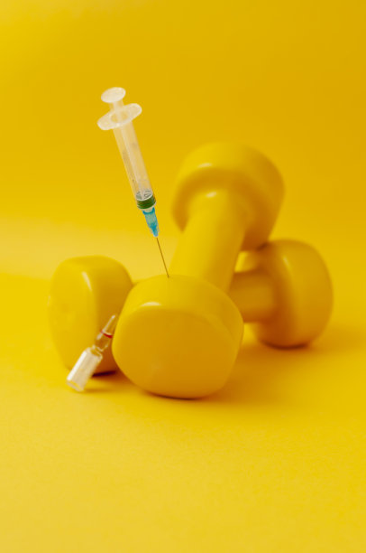 注射器插在一个哑铃里，旁边放着一个医疗安瓿，黄色背景，一张垂直快照。在体育运动中使用兴奋剂的概念，类固醇、睾酮和其他在体育运动中禁用的药物。图片下载