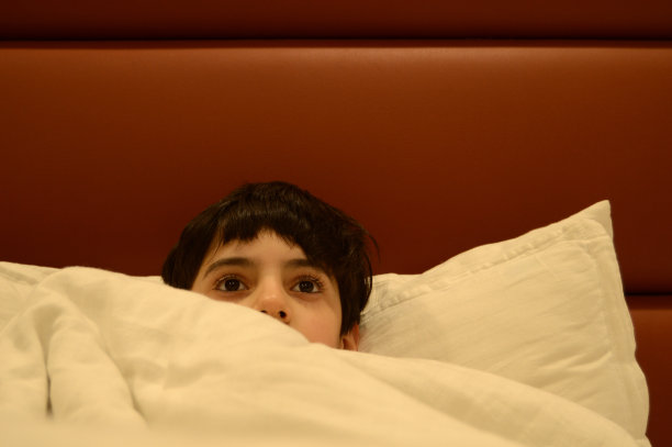 一个可爱可爱的九岁男孩躺在床上枕着枕头休息，白色的床单和羽绒被或被子盖在他身上，看着空白，眼睛睁得大大的图片下载