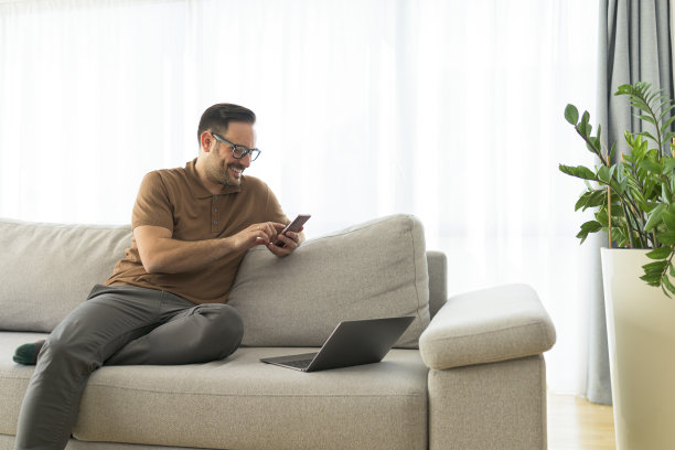 一个英俊开朗快乐的年轻积极的男人在家里的沙发上用手机的形象。图片下载