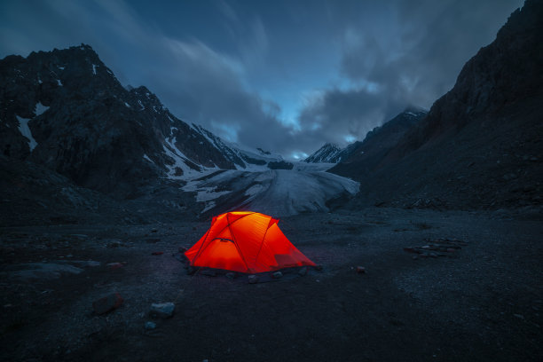 令人敬畏的山地景观与生动的橙色帐篷附近的大冰川舌在夜间星空下的云。帐篷在橘黄色的灯光下闪耀，在星夜中可以看到冰川和山脉的剪影。图片下载