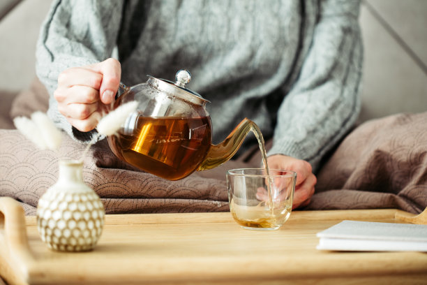 女人从玻璃茶壶中倒茶。在舒适舒适的卧室里吃早餐。图片下载