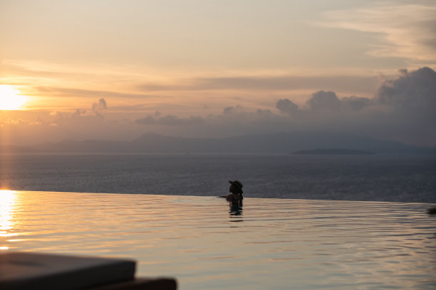 一个戴着帽子的女人在无边的游泳池里放松，看着美丽的日落和海景图片下载