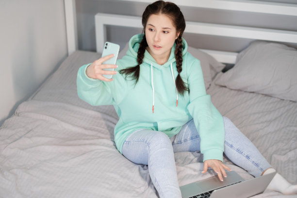 在家工作或学习。年轻的黑发女人，穿着蓝绿色的连帽衫和牛仔裤躺在床上，手里拿着笔记本电脑和蓝绿色的手机。检疫限制。遥远的工作、学习。快乐的远程教育图片下载