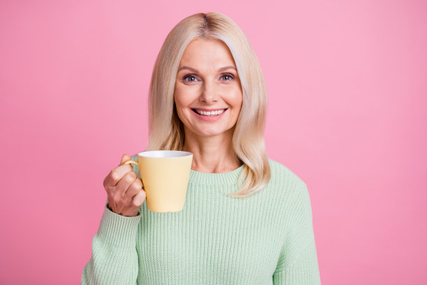 照片肖像的微笑的老妇人在一只手拿黄色的杯子孤立在柔和的粉红色背景图片下载