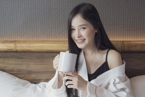 快乐美丽的年轻亚洲妇女放松地喝和看着一杯热咖啡或茶在早上的床上。图片下载