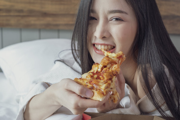快乐的年轻女子在酒店房间的床上吃披萨喝香槟。图片下载
