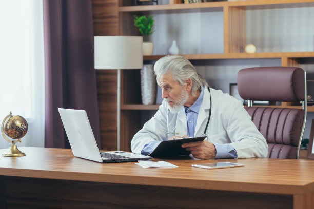 白发苍苍、经验丰富的男医生坐在办公室里，用笔记本电脑远程指导病人图片下载