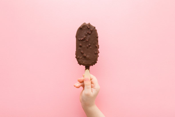 小女孩手拿着带坚果和巧克力釉的冰淇淋，放在浅粉色的桌子背景上。柔和的颜色。特写镜头。孩子们在夏天吃冷的甜食。自顶向下的观点。图片下载