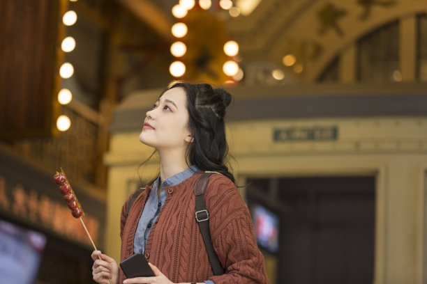 年轻的女大学生拿着手机和糖葫芦在一个老的步行街股票照片图片下载