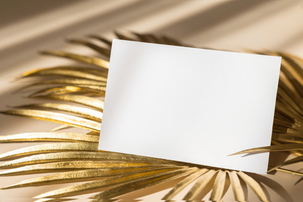 邀请卡模型与金黄色棕榈叶在米色柔和的背景。俯视图，平躺，复制空间。模板空白的白纸模拟为品牌和广告图片下载