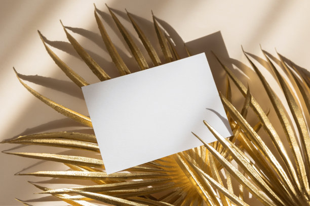 邀请卡模型与金黄色棕榈叶在米色柔和的背景。俯视图，平躺，复制空间。模板空白的白纸模拟为品牌和广告图片下载