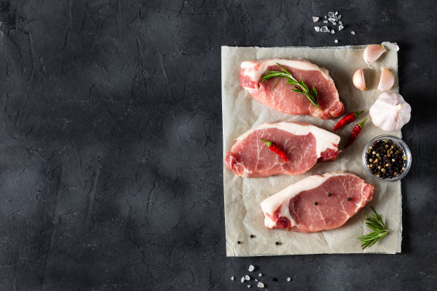一片生的新鲜猪肉，加上迷迭香和大蒜，放在黑色背景的蜡纸上。肉和香料图片下载
