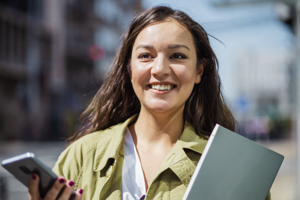 一名年轻女子在使用手机时手持笔记本电脑的肖像图片下载
