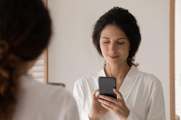 西班牙妇女穿着浴袍站在浴室里拿着智能手机图片下载