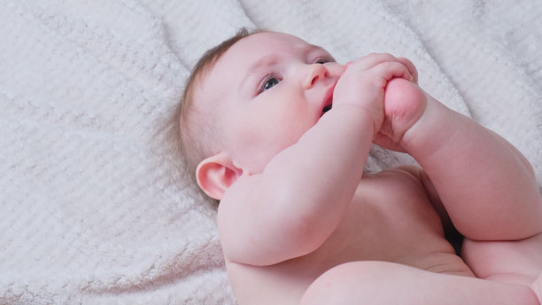 一个快乐的婴儿躺在床上，把他的腿放进嘴里，八个月大。沙发上笑着的蹒跚学步的孩子，近距离看图片下载
